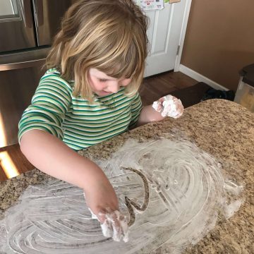 Child writing the letter m in shaving cream on kitchen counter.